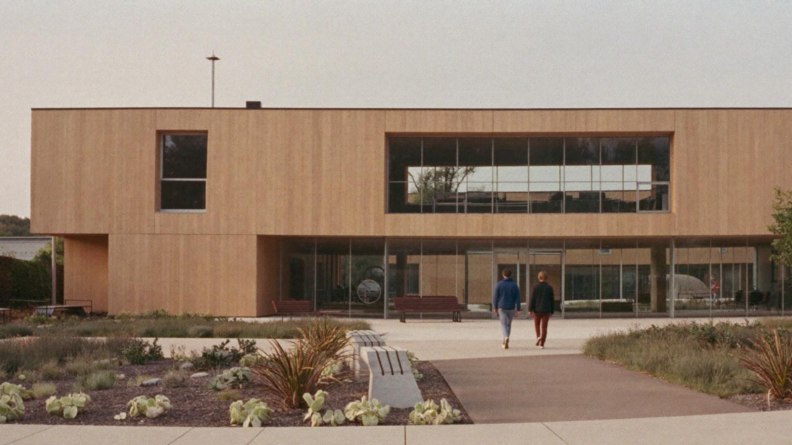A contemporary timber-clad community centre with tall glass, a landscaped forecourt and two people walking toward the entry.