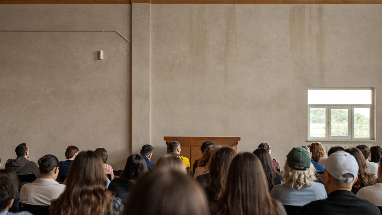 Residents seated at an indoor community information session, seen from behind and facing a bare wall and a lectern.