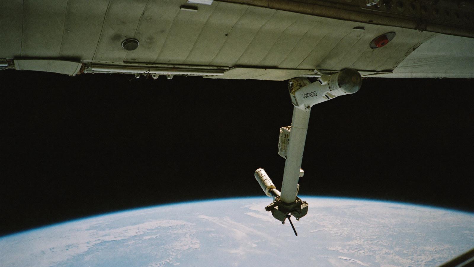 A robotic arm extends from a spacecraft above a blue planet.