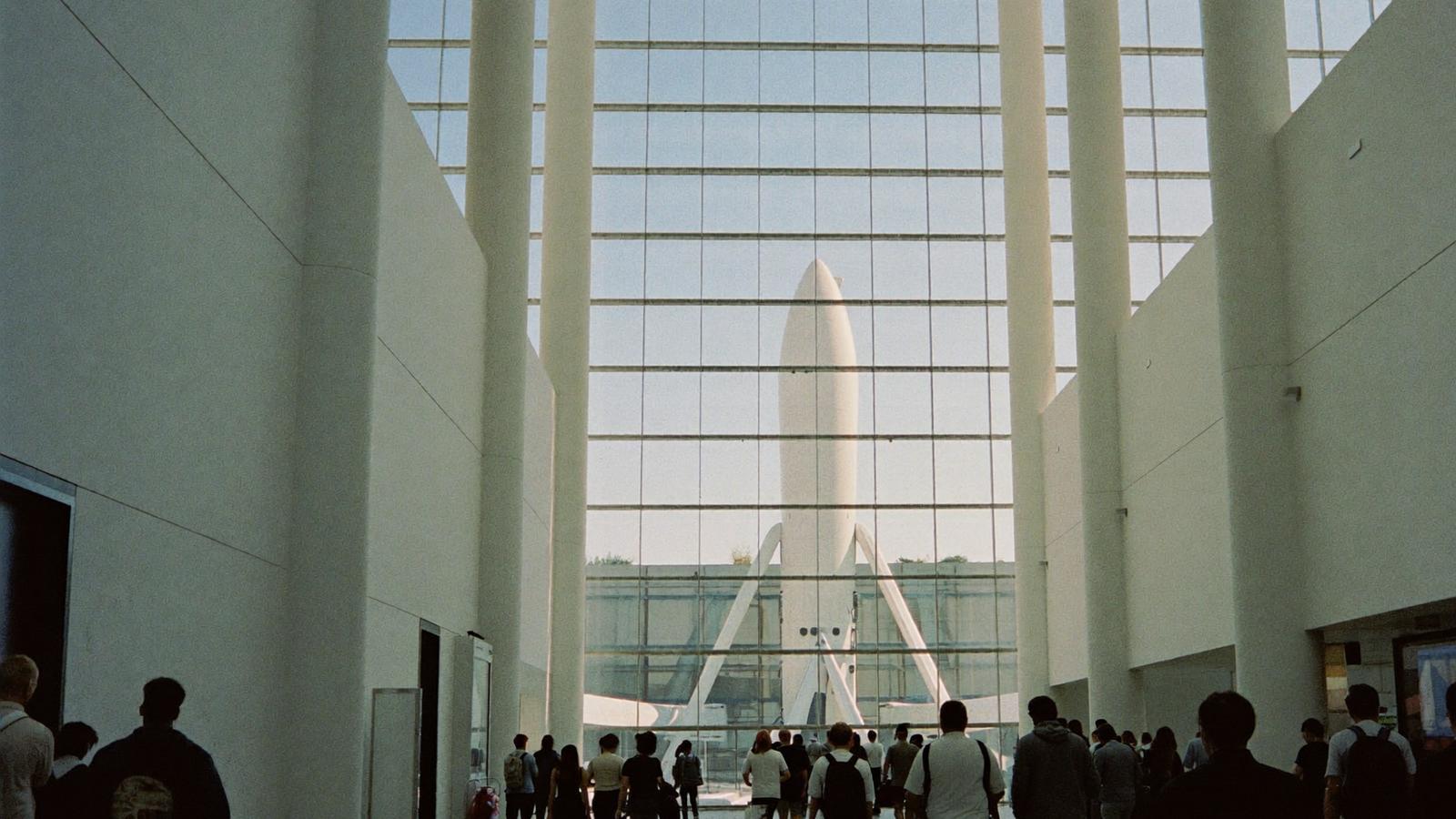 Travellers with luggage crossing a bright terminal concourse toward a glass wall, with a launch vehicle standing on the apron outside.